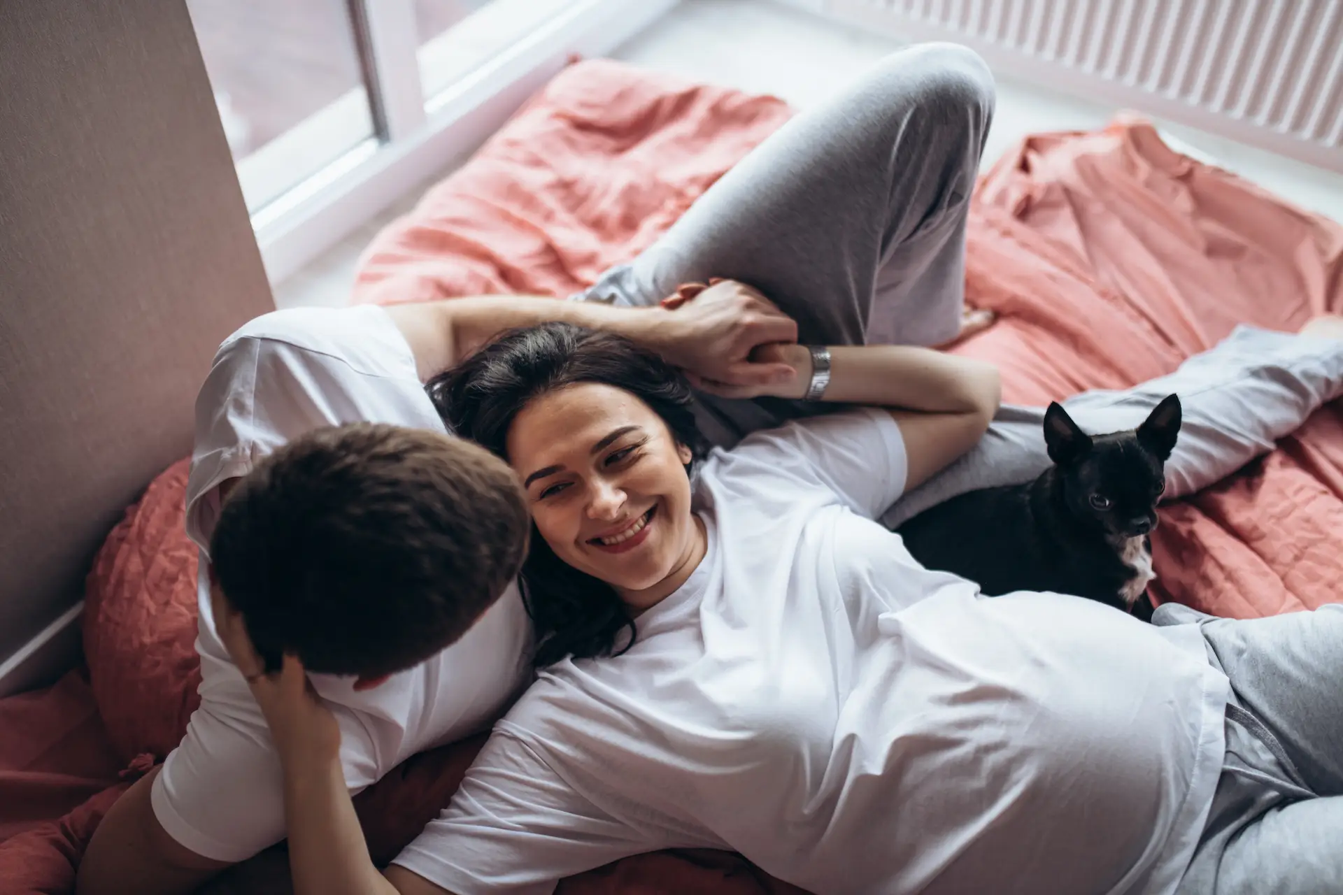 Soft focus. Pregnant woman, man and dog lying on a bed near the window. Attractive couple look to each other and smiling. Natural light, terracotta bed linen. close up shot.