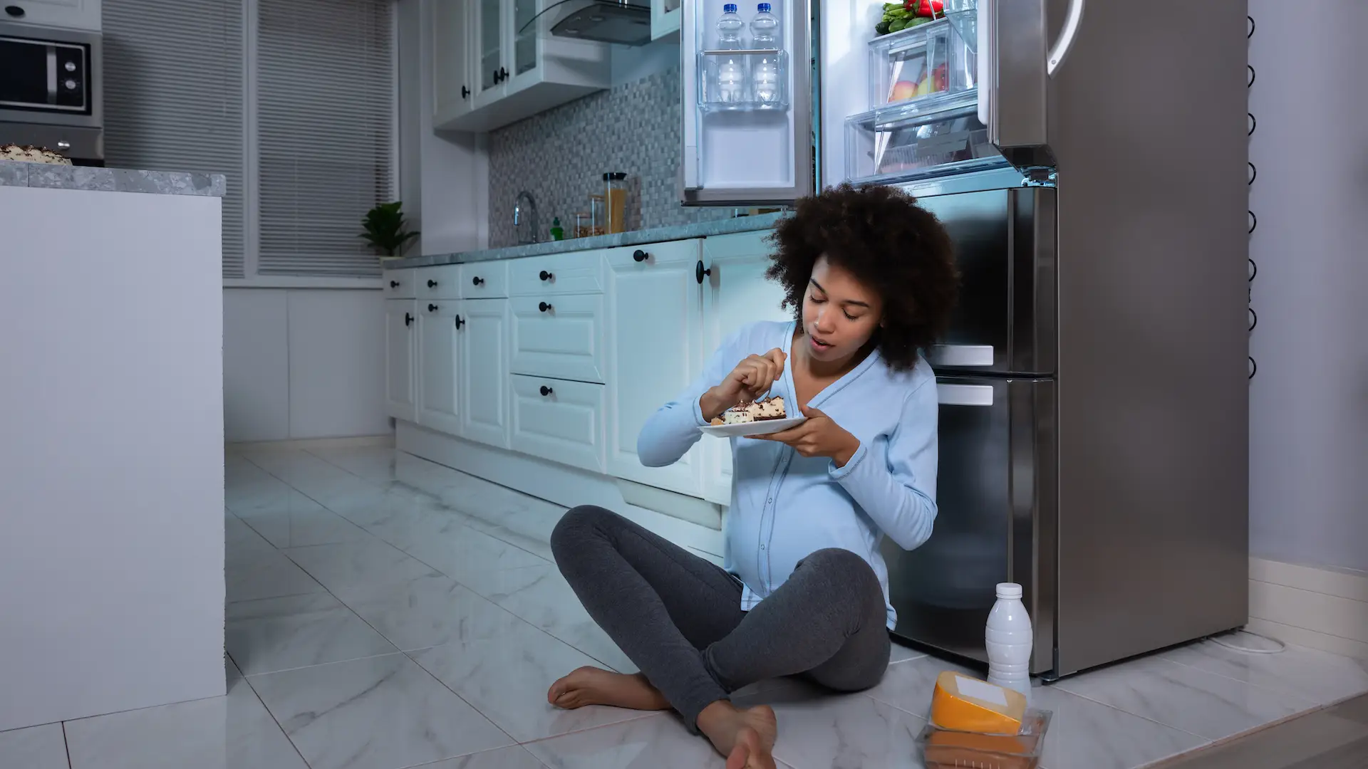 Young Pregnant Woman Eating Piece Of Cake Sitting In Front Of Open Refrigerator At Kitchen