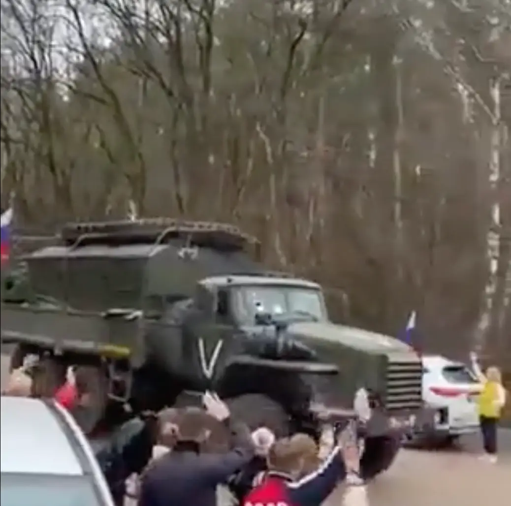 A Russian military convoy with a white painted symbol on its side, being waved into battle by passersby.